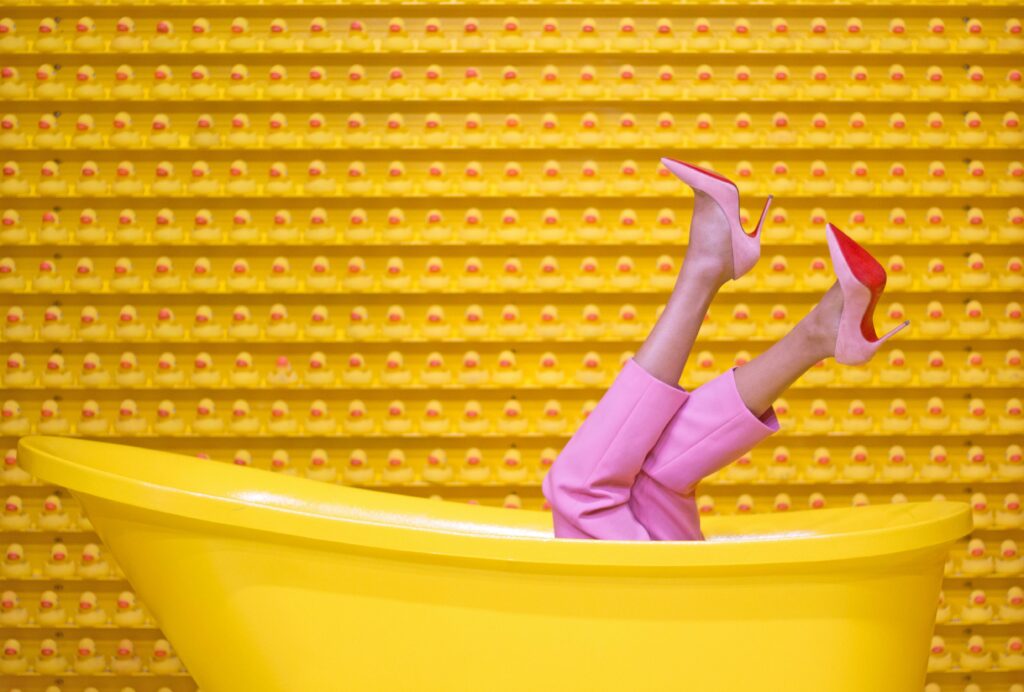 Legs in pink pants and red high heels pop out of a yellow bathtub against a creative rubber duck wall.